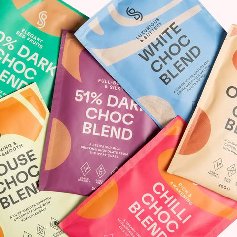 A colorful assortment of Blendsmiths drinking chocolate sachets, featuring flavors like 51% Dark Choc Blend, White Choc Blend, Chilli Choc Blend, and more. Each sachet has a modern design with unique color schemes and highlights key attributes such as veg