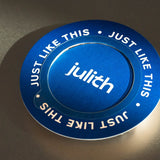 A circular metallic blue product with white text reading 'JUST LIKE THIS' and the brand name 'julith' in the center, photographed on a dark reflective surface.