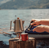 NoNormal Outdoor Coffee Classic Black coffee paste being prepared outdoors beside a camping coffee pot and mug by a lakeside