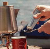 NoNormal Sweet Black Outdoor Coffee paste being squeezed onto a spoon beside a camping coffee pot and mug