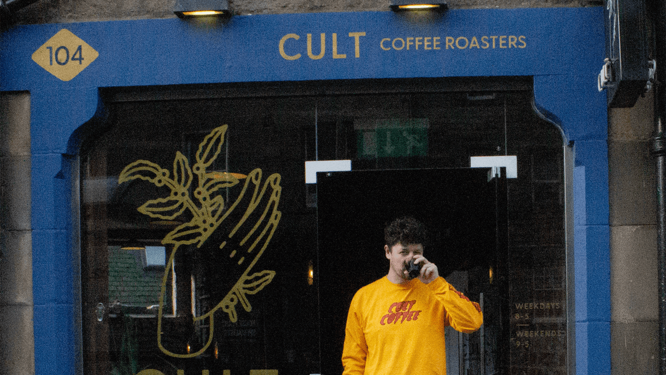 A person in a yellow sweatshirt holds a coffee cup outside Cult Coffee Roasters storefront.