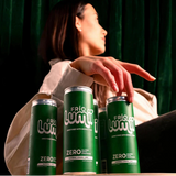 Three green Frigo Lumi Zero energy drinks are lined up in the foreground, with a woman in a light-colored top seated behind them against a dark green curtain background.