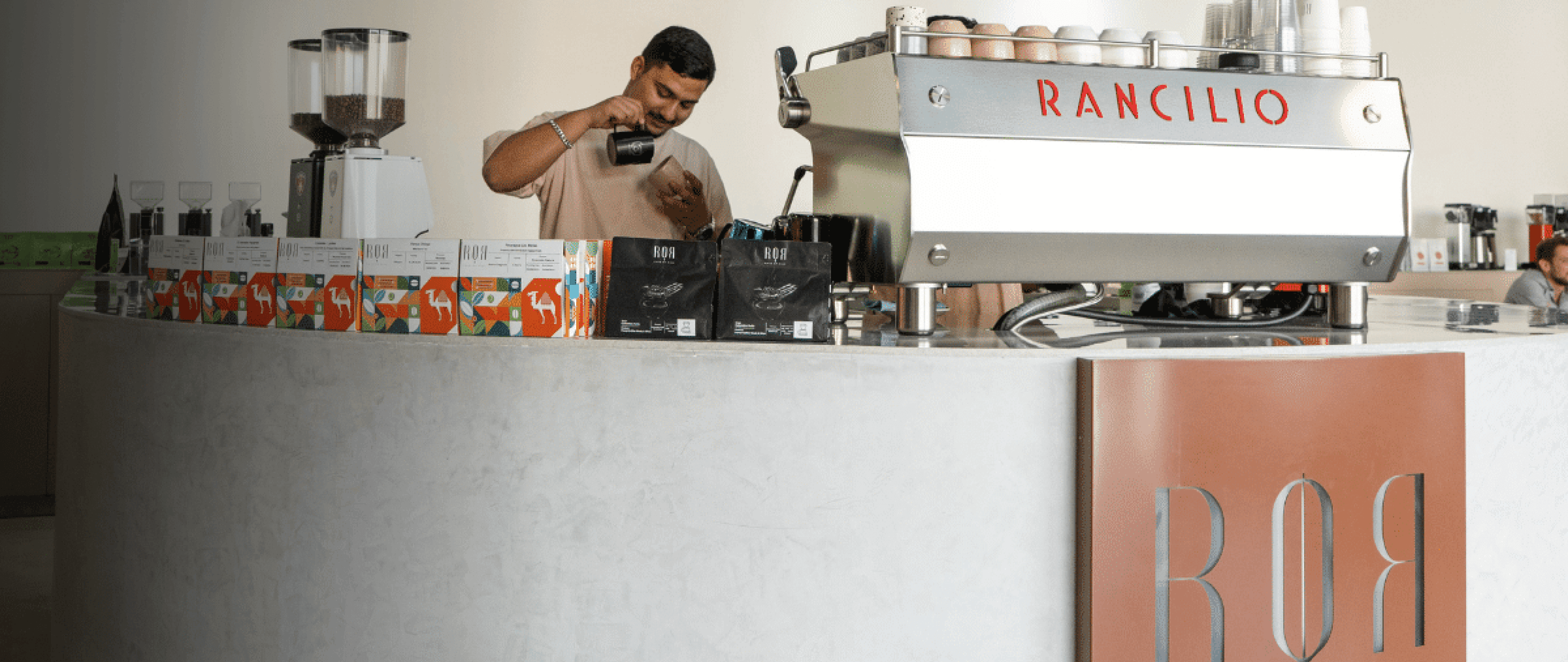 Barista preparing coffee at Rancilio espresso machine in a modern café setting with vibrant packaging.