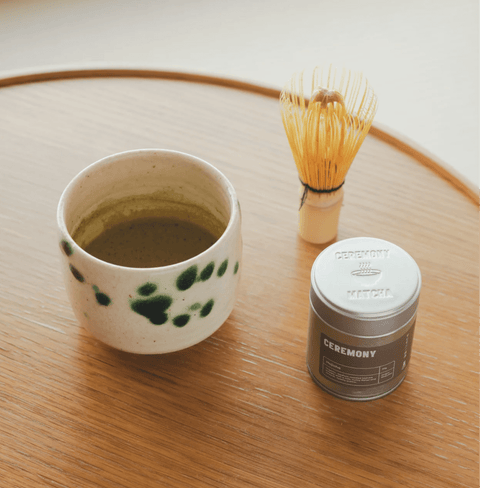 Hojicha tea setup with a ceramic cup, whisk, and tin on a wooden table, showcasing the warm, nutty flavor of roasted Japanese green tea.