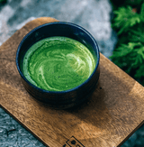 Pure Zen Premium Organic Japanese Matcha Green Tea Powder in a bowl on a wooden plate surrounded by greenery.