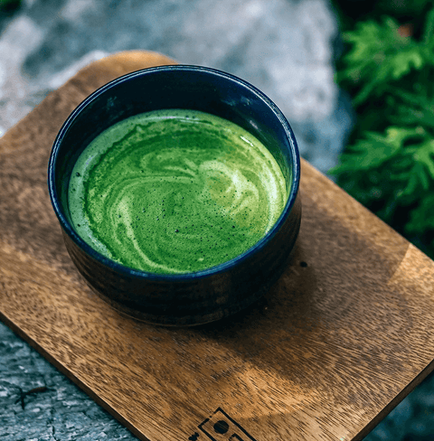 Pure Zen Premium Organic Japanese Matcha Green Tea Powder in a bowl on a wooden plate surrounded by greenery.