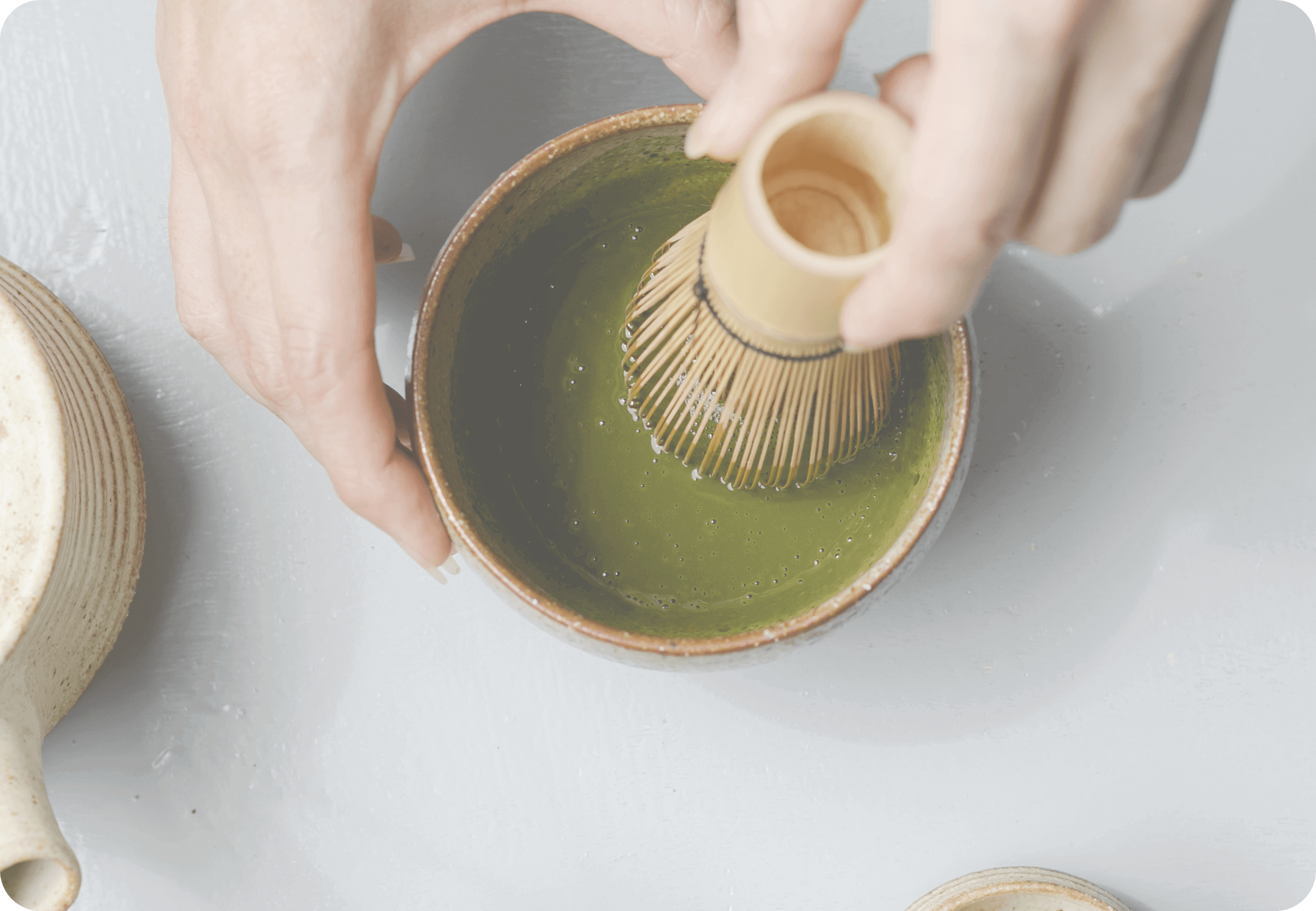 Hands whisking matcha tea in a ceramic bowl with a bamboo chasen on a light tabletop
