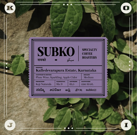 A square product label for SUBKO Specialty Coffee Roasters is centered over a background of green leaves and a stone wall. The label is lavender-purple with black text detailing the coffee origin as Kalledevapura Estate, Karnataka, and roast profile as Medium.