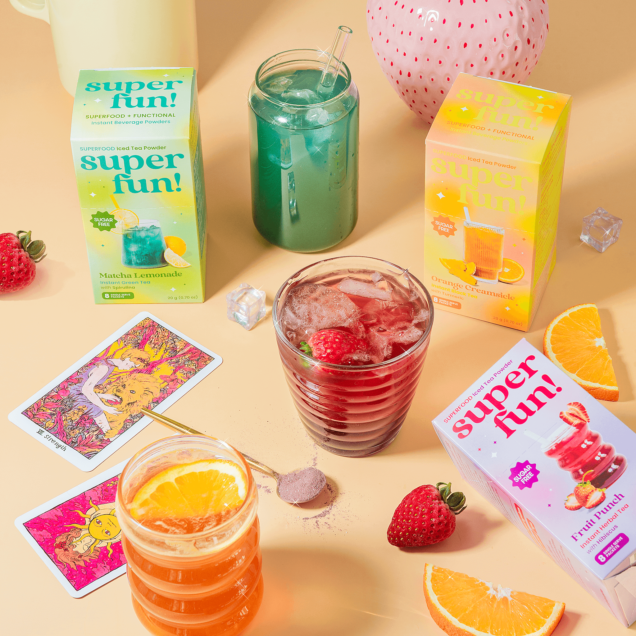 Colorful Superfun beverage boxes with iced fruit drinks, tarot cards, strawberries, and orange slices on a pastel tabletop