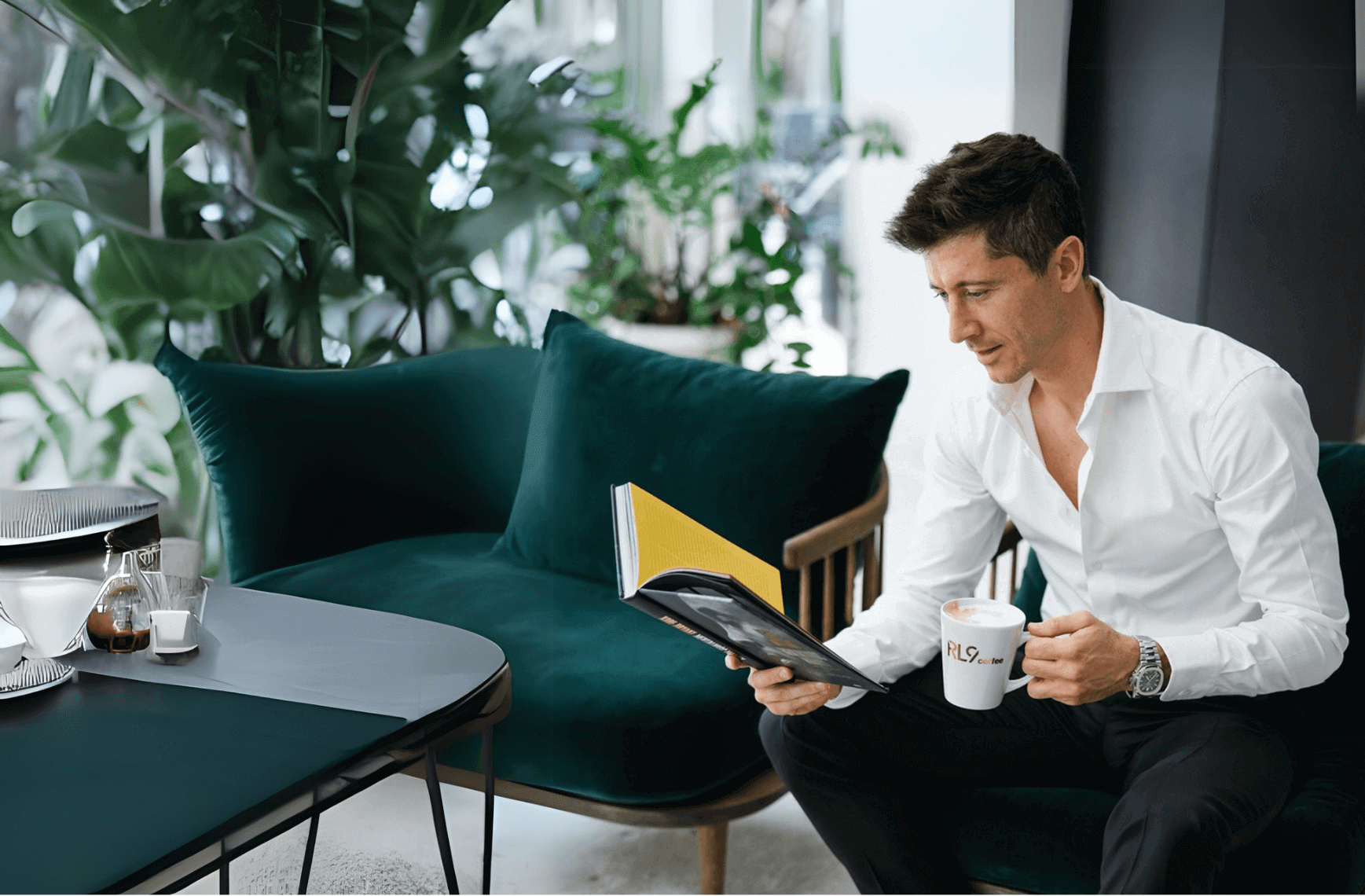 Man in a white shirt enjoying coffee and reading a magazine in a stylish, modern living room with green plants.