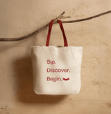 Tote Bag with red handles featuring 'Sip. Discover. Begin.' text on a natural fabric background.