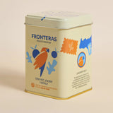A square metal tin of Fronteras Organic Tea, featuring a cream-colored body with colorful illustrations of a parrot, blue leaves, and a stamp, set against a solid light beige background.