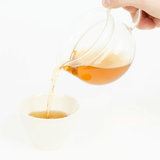 Pouring Chamomile Hojicha tea from a glass teapot into a white tea cup.