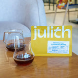 A yellow pouch of Julith coffee on a wooden table, with two glass cups of brewed coffee in front of it and a blurred indoor background.