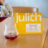 Person pouring a drink into a glass next to a 'julitch' branded box on a wooden surface.