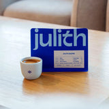 A blue coffee bag package labeled 'julith' stands on a light wood table next to a small white ceramic cup of espresso with crema.