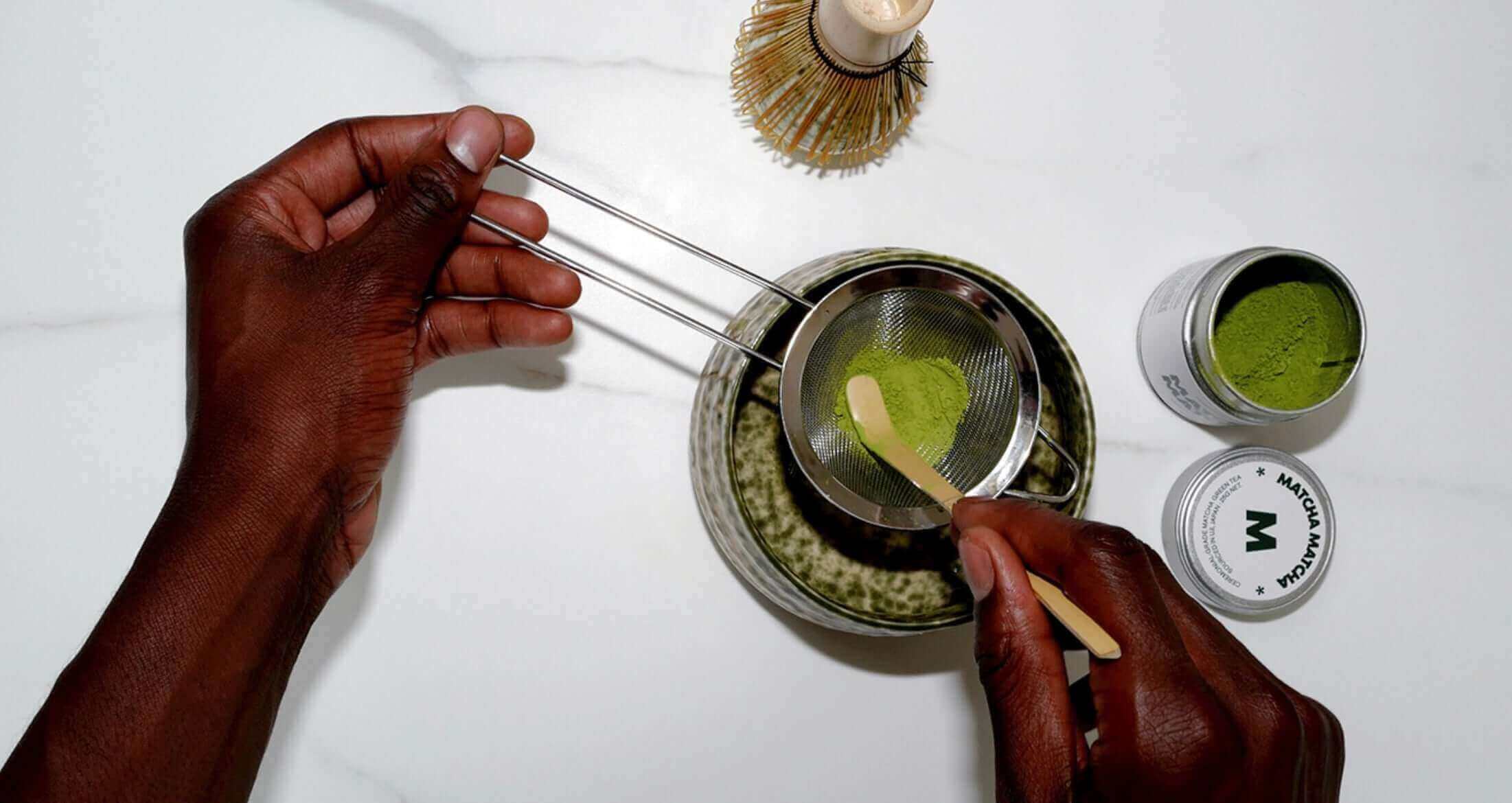 Hands whisking green matcha tea powder in a bowl with a bamboo whisk and matcha tins on a white surface