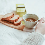 A hand holding a green bowl of Mate Earl Grey Tea Bags with sliced bread on a wooden board.