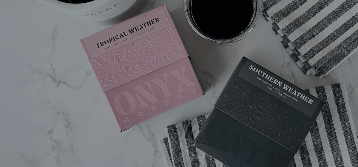 Pink Tropical Weather and dark Southern Weather OMYF coffee boxes displayed on marble with striped cloth and coffee cups
