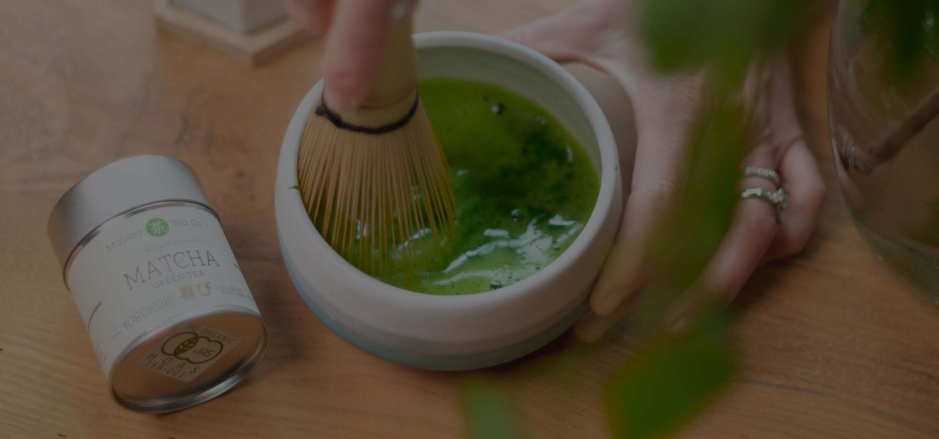 Hands whisking bright green matcha powder in a bowl beside a matcha tin on a wooden table
