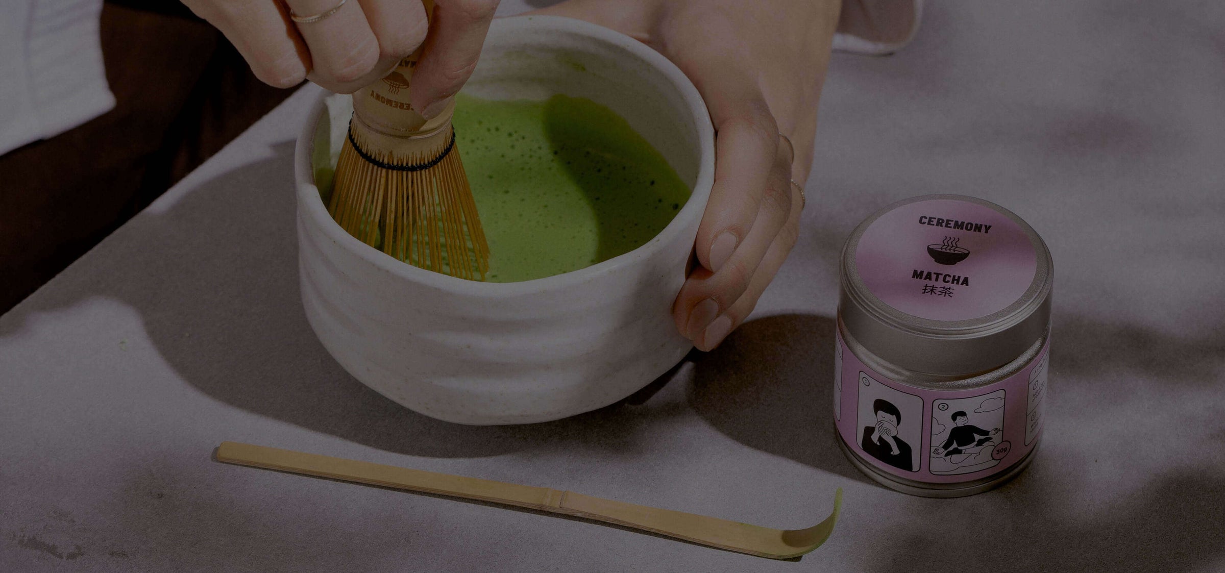 Hand whisking matcha tea in a white bowl beside a tin of ceremonial matcha powder and bamboo whisk