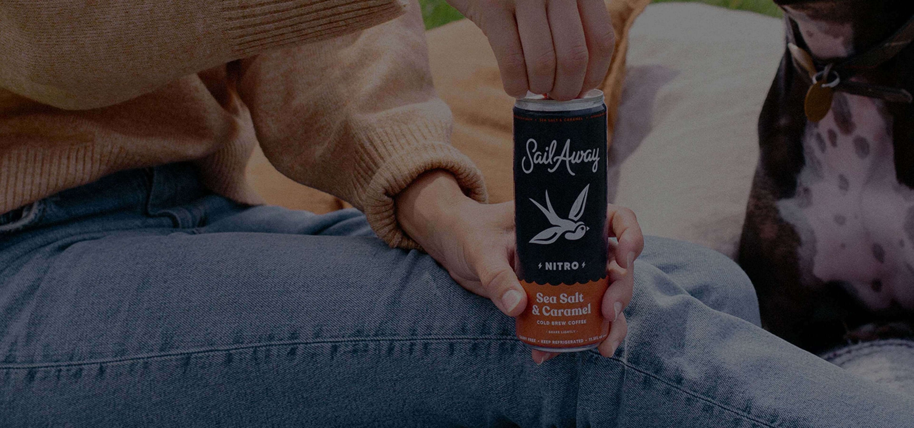 Person holding a Sail Away Nitro Sea Salt Caramel drink can while sitting with a dog